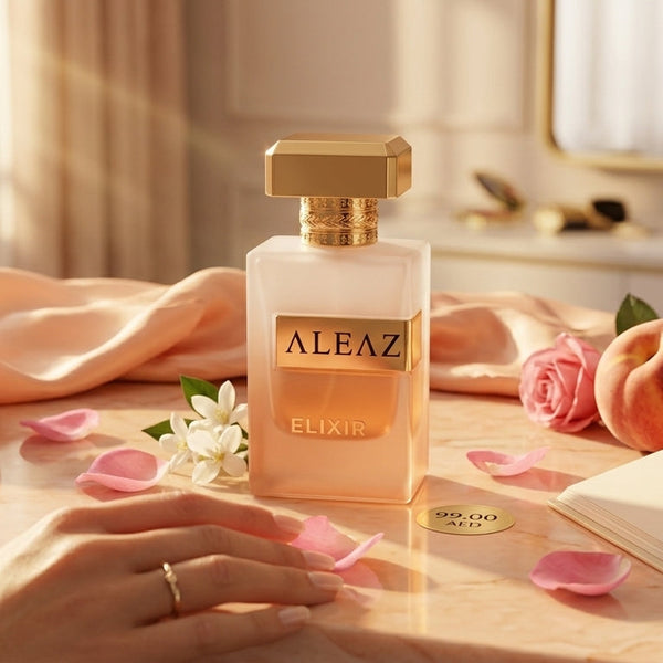 A bottle of ALEAZ Elixir perfume is displayed on a marble tabletop with peach-colored fabric, peaches, pink petals, and an open book in a warm, elegant room setting. A hand with a gold ring rests near the bottle.