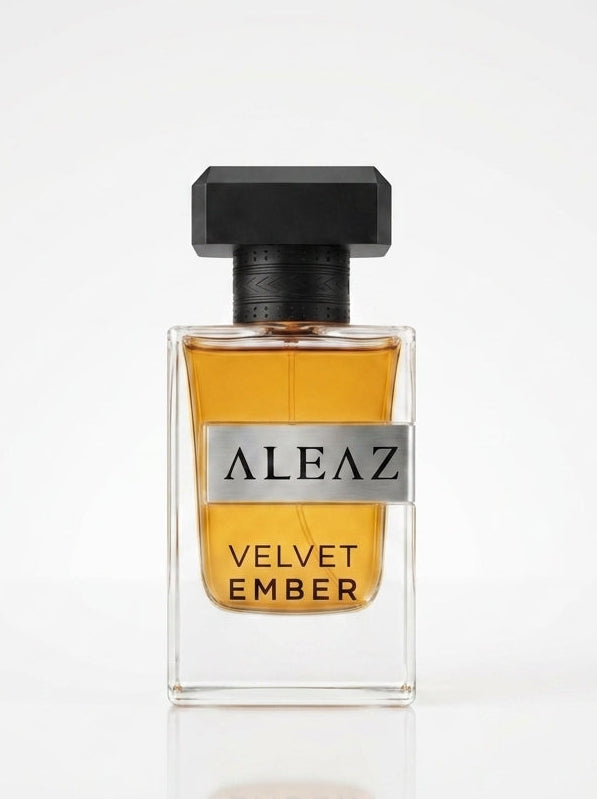A clear rectangular perfume bottle filled with amber-colored liquid, featuring a black cap and a silver label that reads 'ALEAZ VELVET EMBER'. The bottle is centered on a reflective white surface against a clean white background.