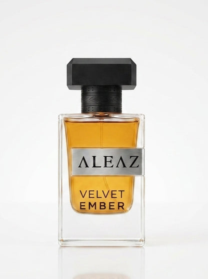 A clear rectangular perfume bottle filled with amber-colored liquid, featuring a black cap and a silver label that reads 'ALEAZ VELVET EMBER'. The bottle is centered on a reflective white surface against a clean white background.